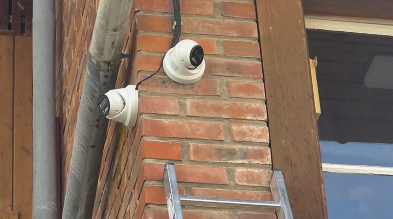 Zwei weisse Hikvision Dome-Kameras an einer Backstein-Hausecke unter Holz-Vordach, sichtbares Aufputz-Kabel