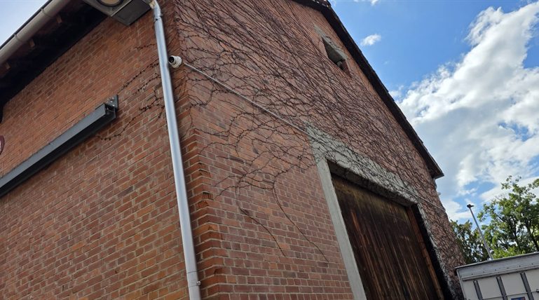 Weisse Hikvision Dome-Kamera an der oberen Ecke eines Backstein-Giebels, Aufputz-Kabel entlang der Fassade