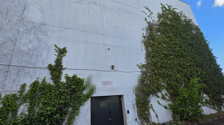 Weisse, efeubewachsene Industriehalle mit einer Kamera über dem Anlieferungstor, Kabel sichtbar verlegt