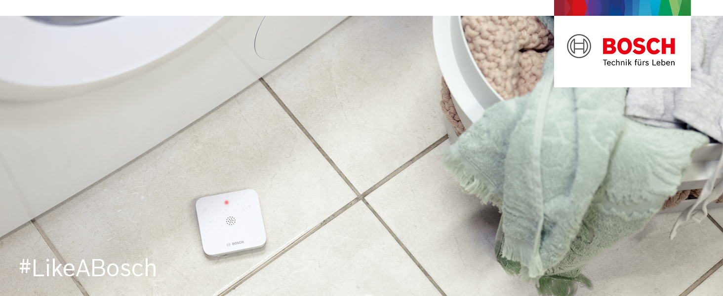 Flacher weisser Bosch Smart Home Wassermelder auf dem Boden neben einer Waschmaschine – Detail 1