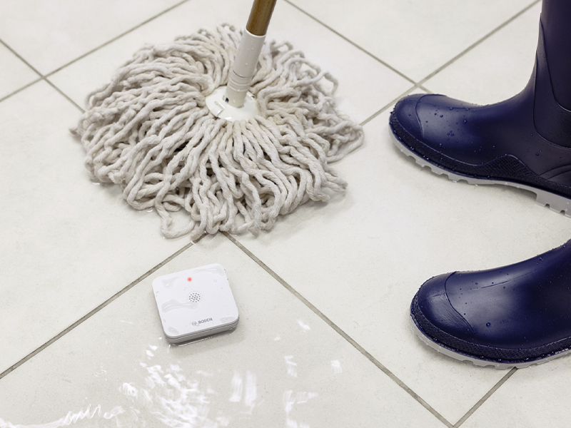 Flacher weisser Bosch Smart Home Wassermelder auf dem Boden neben einer Waschmaschine – Detail 6