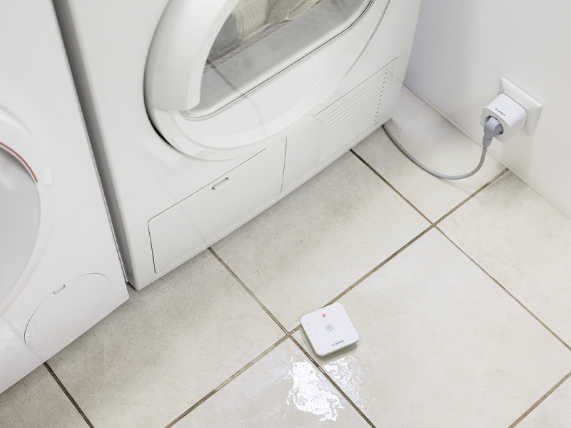 Flacher weisser Bosch Smart Home Wassermelder auf dem Boden neben einer Waschmaschine – Detail 7