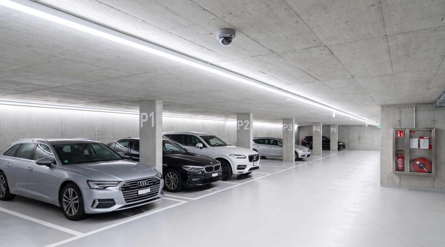 Graue Dome-Kamera an der Betondecke einer hellen modernen Tiefgarage ueber einer Reihe geparkter Autos