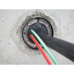 Eingebaute Mauerdurchführung von Ritzer mit Pressplatte aus Edelstahl in einer Kernbohrung, durch die ein schwarzes Rohr und zwei Kabel geführt sind